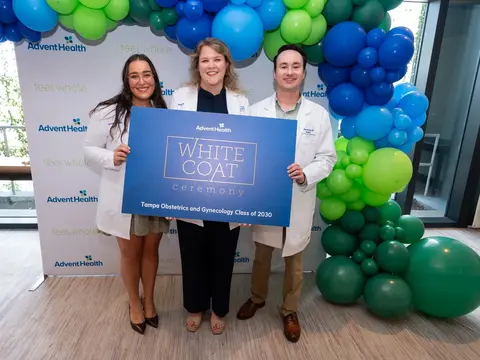 Three people in white coats hold a White Coat Ceremony sign beneath a blue and green balloon arch in front of an AdventHealth backdrop.