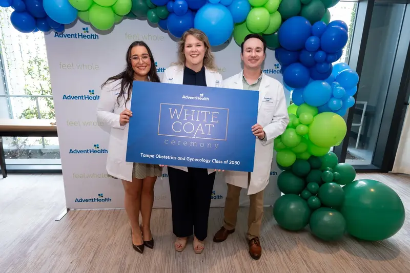 Three people in white coats hold a White Coat Ceremony sign beneath a blue and green balloon arch in front of an AdventHealth backdrop.