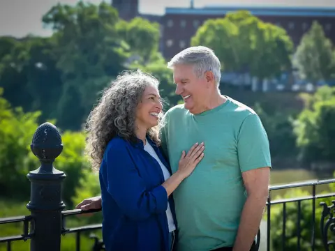Smiling couple standing together on a balcony overlooking a river and park with lush green trees and historic buildings in the background