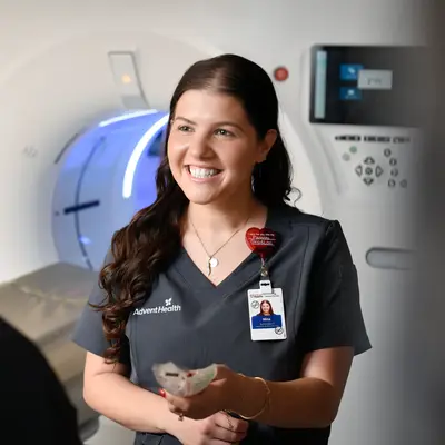 Smiling female AdventHealth team member in navy scrubs holding paperwork while standing in front of an illuminated medical imaging scanner in a clinical setting
