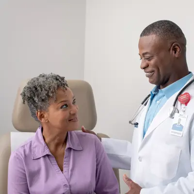 A smiling doctor in a white coat with a stethoscope around his neck stands next to a smiling patient in a purple shirt.