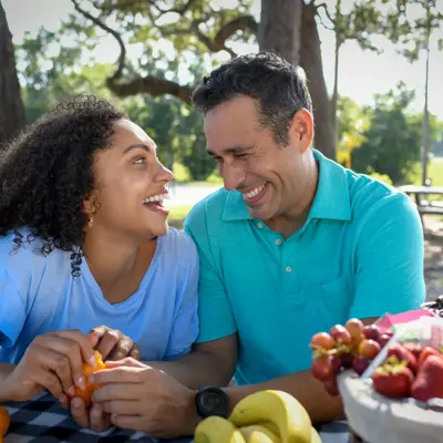 A smiling man and woman share a picnic, enjoying fruit and conversation in a park.
