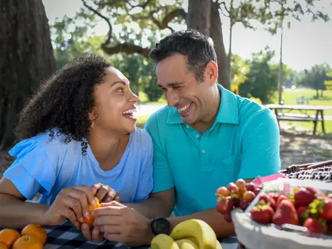 A smiling man and woman share a picnic, enjoying fruit and conversation in a park.