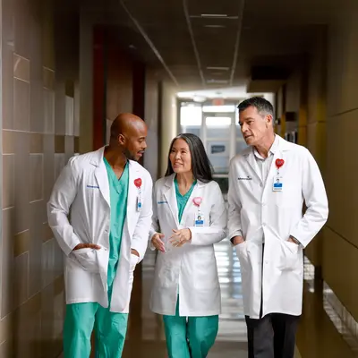 Three healthcare professionals in white coats and green scrubs walking down a hospital corridor.