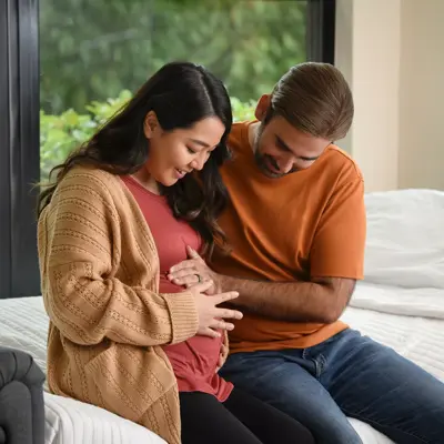 A pregnant woman and her partner sit together on a bed, smiling and holding her belly.