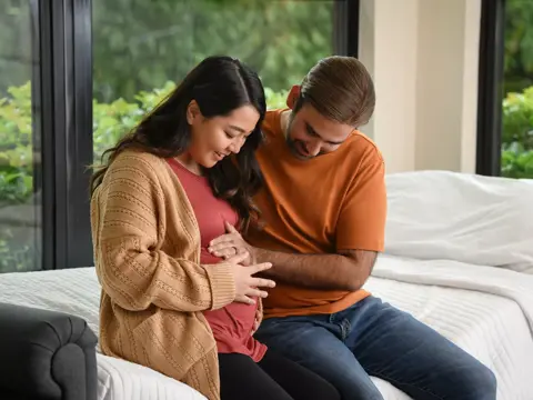 A pregnant woman and her partner sit together on a bed, smiling and holding her belly.