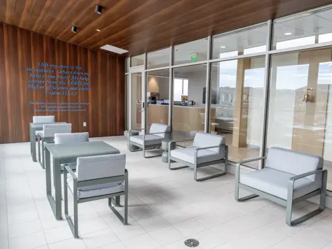 Outdoor healing terrace with modern seating arrangements featuring light gray cushioned chairs, warm wood-paneled ceiling, floor-to-ceiling glass walls, and an inspirational biblical quote displayed on the wooden wall