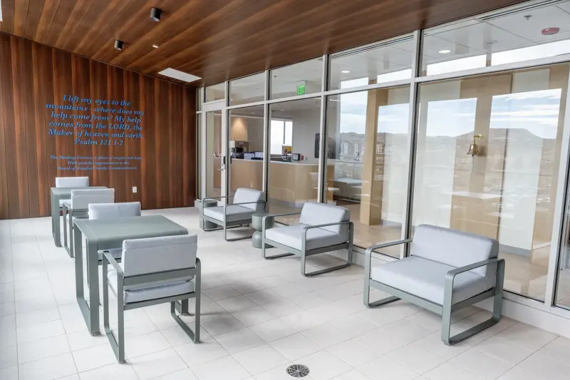 Outdoor healing terrace with modern seating arrangements featuring light gray cushioned chairs, warm wood-paneled ceiling, floor-to-ceiling glass walls, and an inspirational biblical quote displayed on the wooden wall