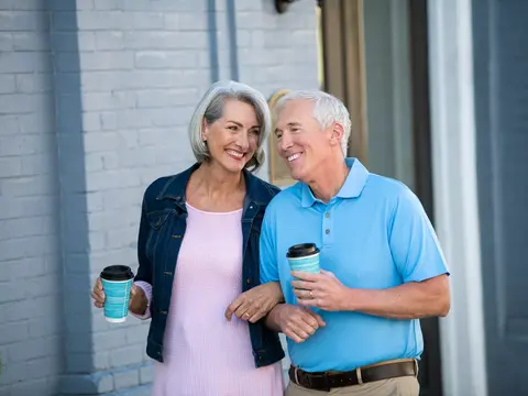 Elderly couple smiling and holding coffee cups outside a building.