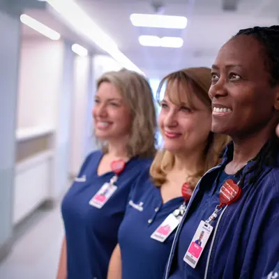 Four smiling nurses in blue uniforms stand together in a hospital hallway.