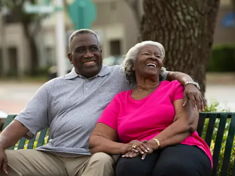 Elderly couple sitting on a bench, smiling and embracing each other in a park.