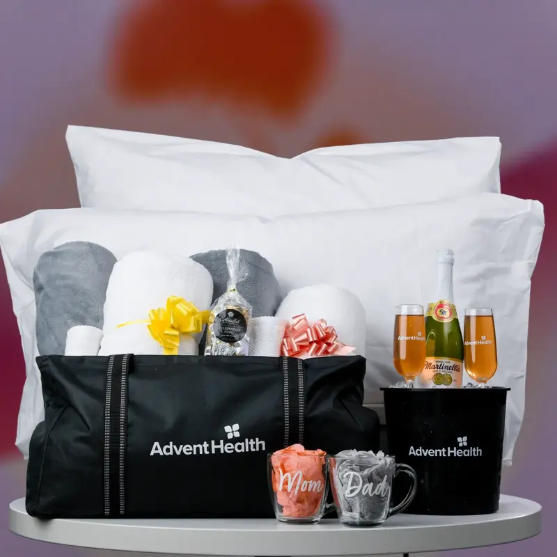 Gift package display featuring two black AdventHealth tote bags filled with white pillows and blankets, decorative bows, Martinelli's sparkling cider with champagne flutes, chocolates, and Mom and Dad mugs arranged on a white round table