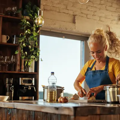 Two adults cook at a kitchen island, chopping vegetables beside a pot; sunlight, plants, shelves in background.