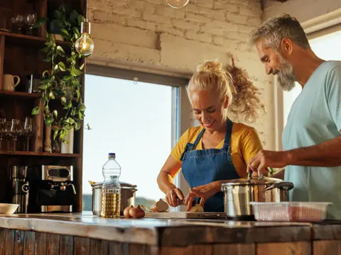 Two adults cook at a kitchen island, chopping vegetables beside a pot; sunlight, plants, shelves in background.