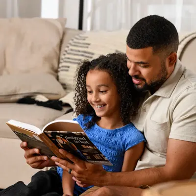 Father and daughter reading a book together while sitting on a couch in a bright, modern living room