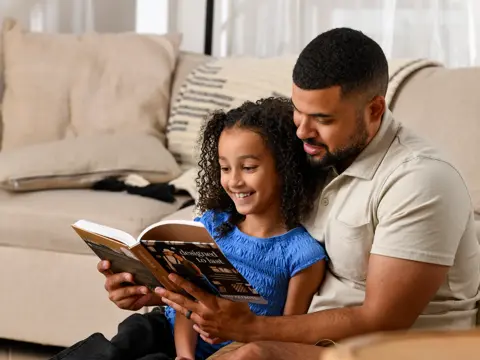 Father and daughter reading a book together while sitting on a couch in a bright, modern living room