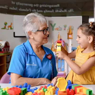 An older woman and a young girl are playing with colorful building blocks at a table in a playroom.