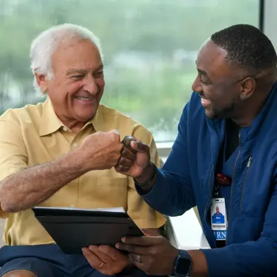 Two smiling men, one elderly and one younger, engaging in a friendly handshake while looking at a tablet.