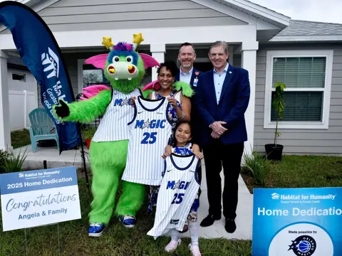 A family holding up Orlando magic gear in front of a house with AdventHealth Officials.