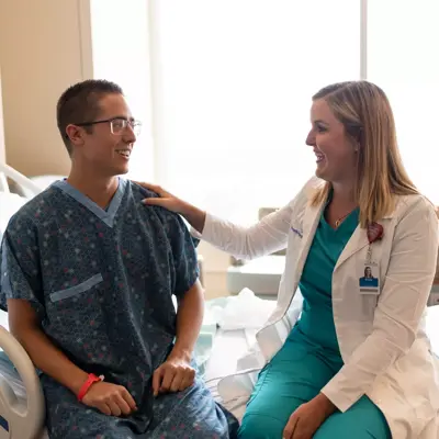 Female doctor gently touches the shoulder of her male patient as they sit on the side of a hospital bed.