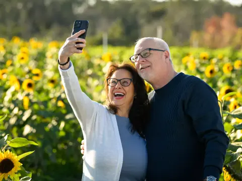 Elderly couple smiling and taking a selfie in a sunflower field.
