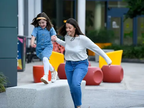 Mother and daughter holding hands, jumping on a concrete bench in a colorful outdoor area.