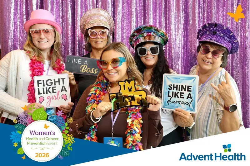 Five women wearing colorful hats and glasses pose with empowering signs at the 2026 AdventHealth Women’s Health and Cancer Prevention Event.