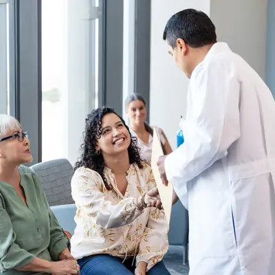 Doctor in white coat hands a document to a smiling seated woman as another woman sits beside her in a waiting area.