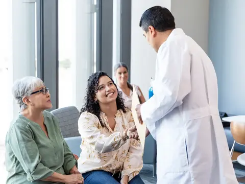 Doctor in white coat hands a document to a smiling seated woman as another woman sits beside her in a waiting area.