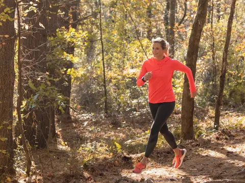 A woman in red shirt and black leggings jogging in a forest with sunlight filtering through the trees.