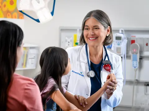 A smiling doctor in a white coat with a stethoscope around her neck is holding the hand of a young girl while talking to her mother in a hospital room.
