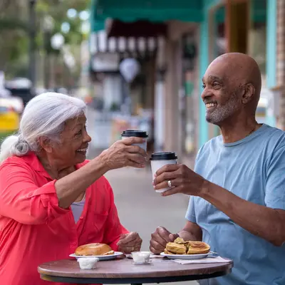 Elderly couple sharing a meal and coffee at an outdoor table, smiling and engaged in conversation.