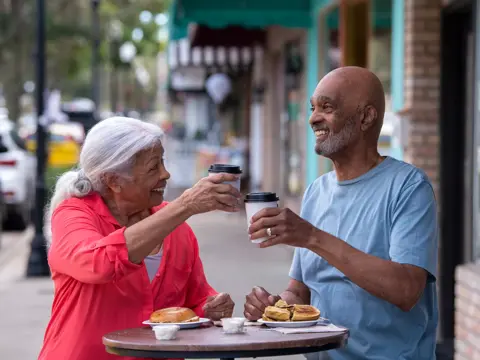 Elderly couple sharing a meal and coffee at an outdoor table, smiling and engaged in conversation.
