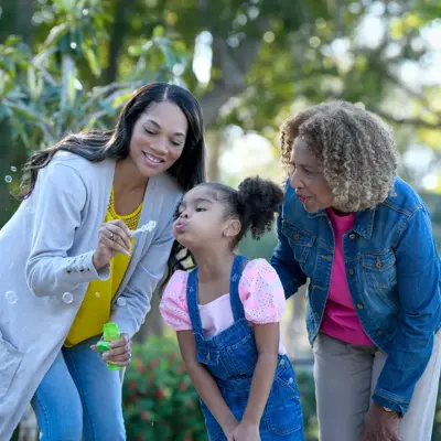 Three generations of women blowing bubbles together in a park, smiling and enjoying the moment.
