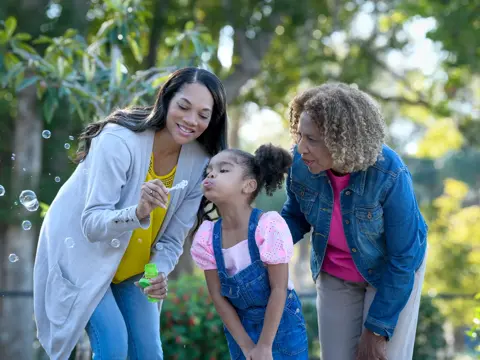 Three generations of women blowing bubbles together in a park, smiling and enjoying the moment.