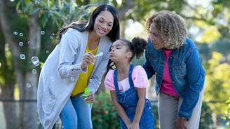 Three generations of women blowing bubbles together in a park, smiling and enjoying the moment.