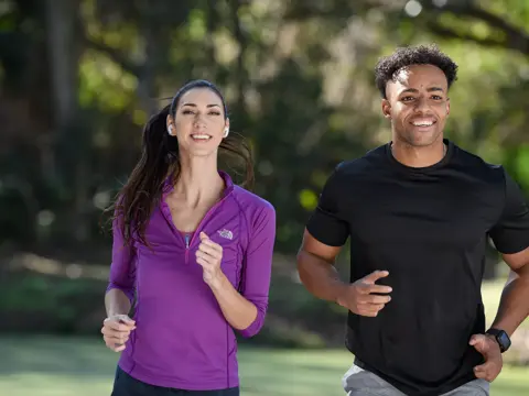 A woman and a man are jogging outside in the park, both smiling and wearing wireless earbuds.