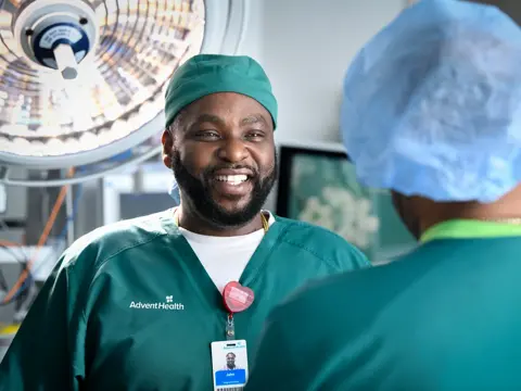 A smiling AdventHealth employee in scrubs stands in an operating room, embodying warmth and hope.