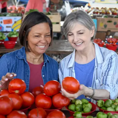 Two women smiling and selecting fresh tomatoes at an outdoor market.