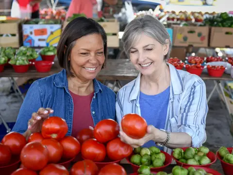 Two women smiling and selecting fresh tomatoes at an outdoor market.