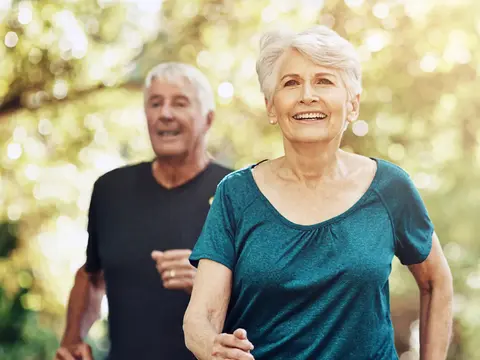 An older couple jogging outdoors on a sunny day, with a smiling woman in a teal shirt in the foreground and a man in a black shirt behind her in a park setting