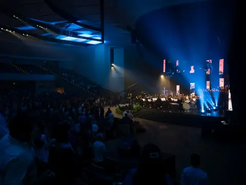 Audience standing in a dark auditorium as an orchestra and choir perform on a blue-lit stage.