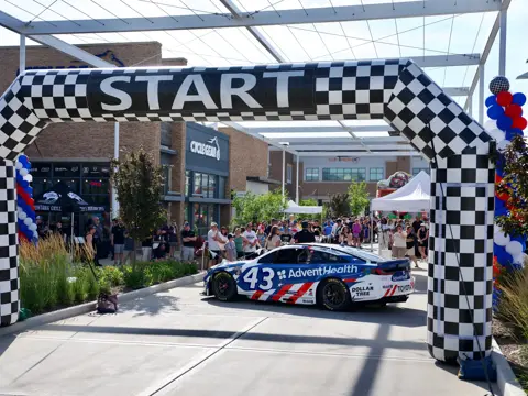 Branded race car sits beneath a checkered start arch as spectators gather at an outdoor event with red, white, and blue balloon columns.