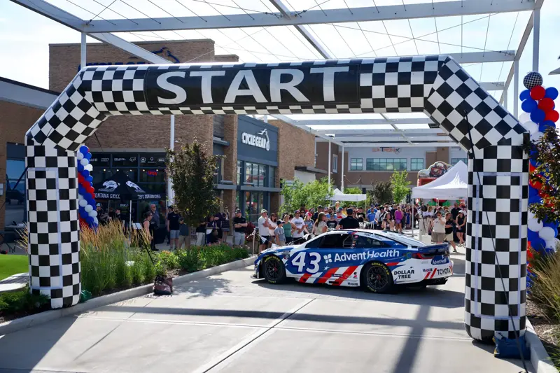 Branded race car sits beneath a checkered start arch as spectators gather at an outdoor event with red, white, and blue balloon columns.