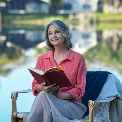 A woman with gray hair sits by a lake, smiling while reading a book.