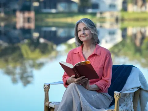 A woman with gray hair sits by a lake, smiling while reading a book.