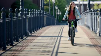 Young woman riding a bicycle on a pedestrian bridge with ornate black railings and vintage street lamps on a sunny day