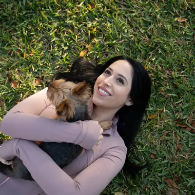A woman with long black hair is lying on the grass, smiling and holding a small dog.