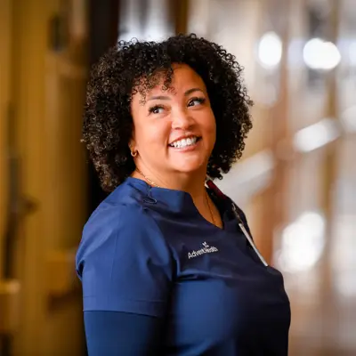 A smiling woman in a blue AdventHealth uniform stands in a hallway, conveying warmth and positivity.