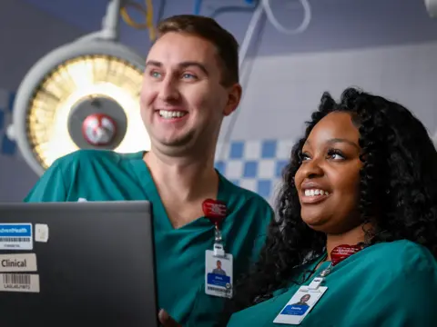 Two smiling healthcare team members in teal scrubs collaborating on a laptop in a clinical setting with surgical lights in the background